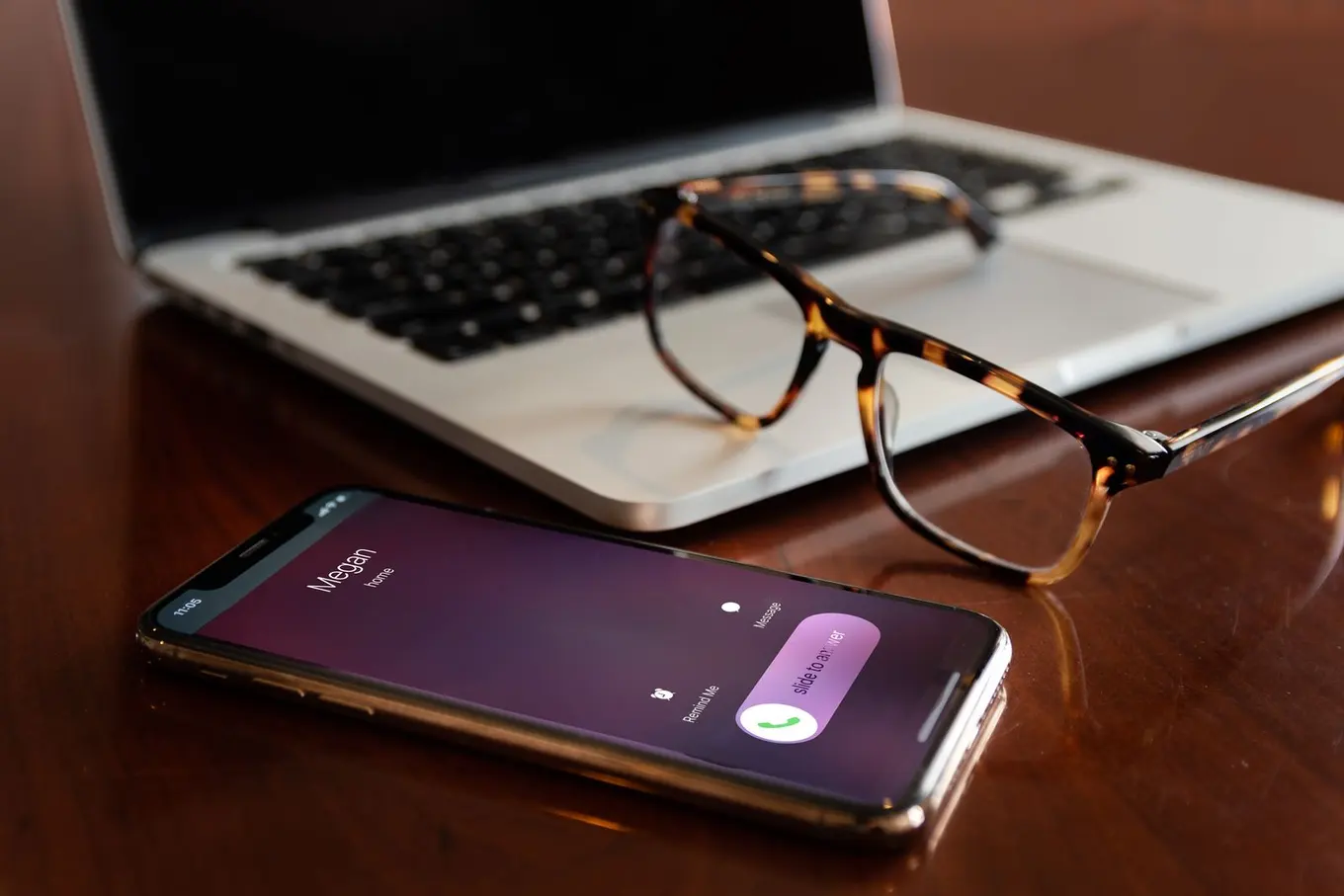 A phone on a wooden desk showing an incoming call, next to a laptop and a pair of glasses.
