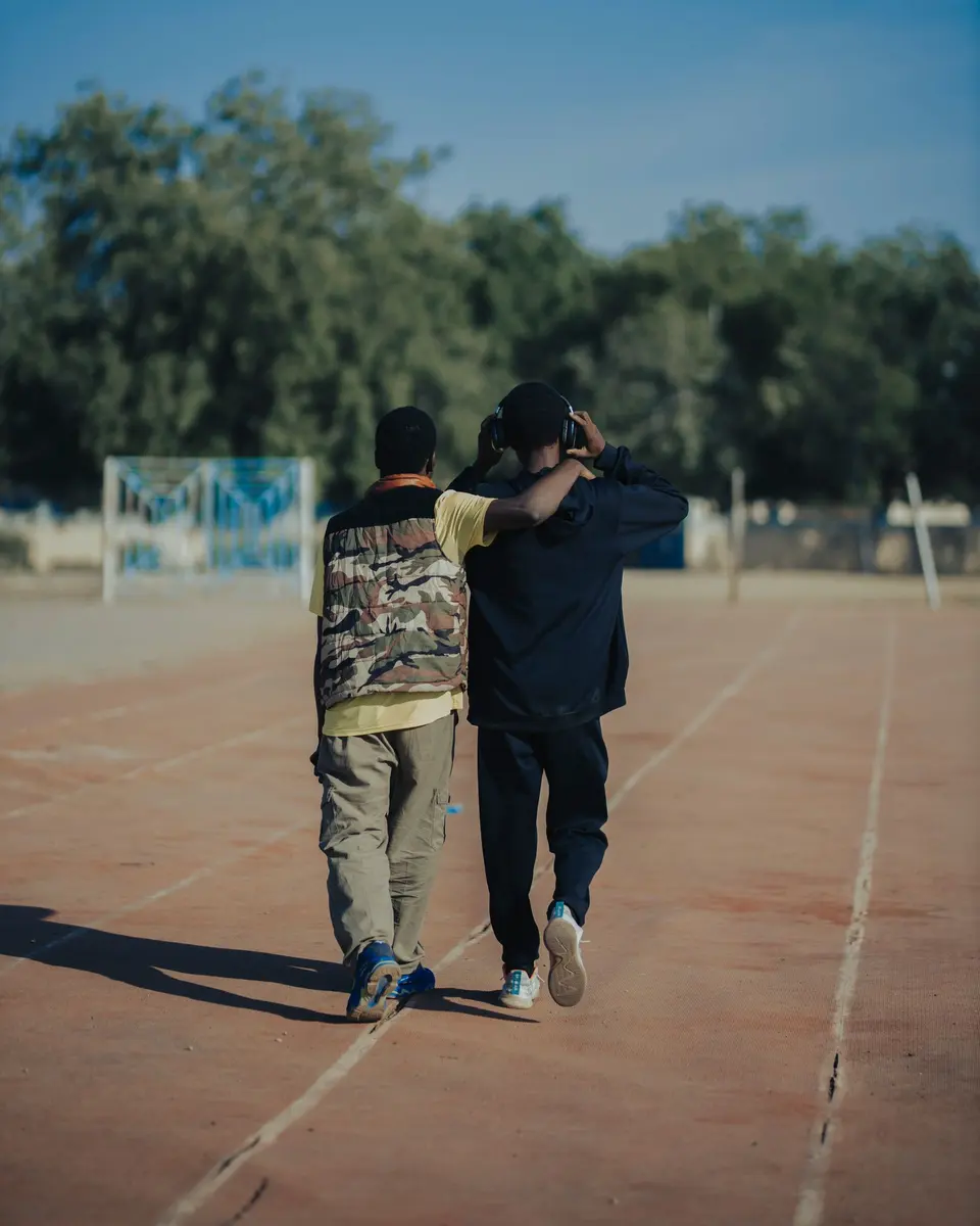 Two friends walking side by side on a running track, one with an arm around the other, seen from behind.