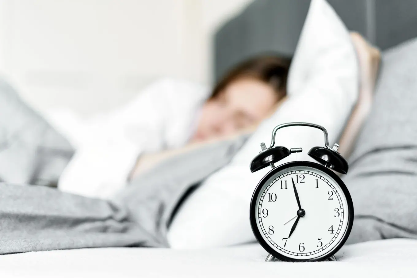 A person asleep in bed behind an analogue alarm clock, face out of focus.