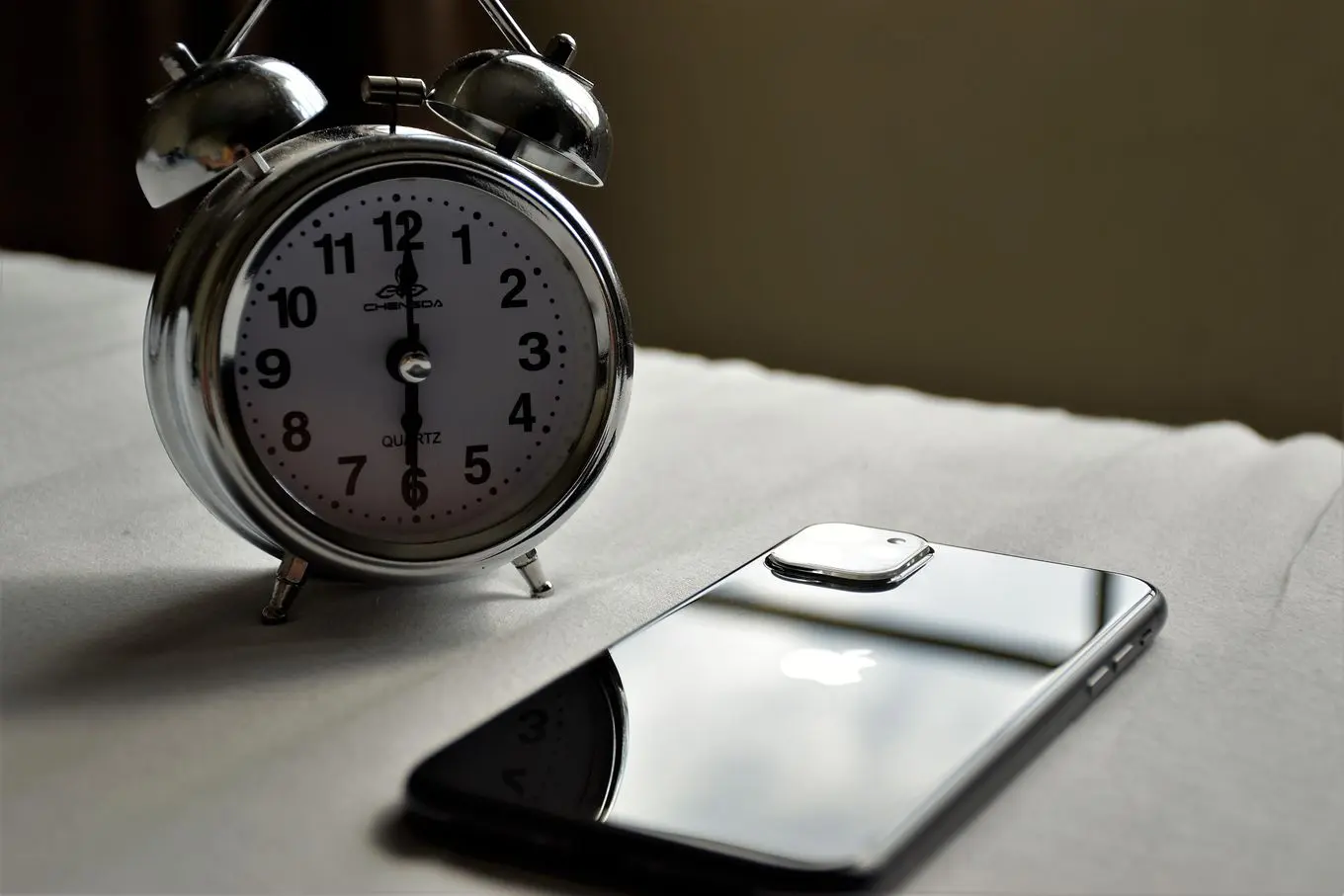 A silver twin-bell alarm clock next to a smartphone on a white surface
