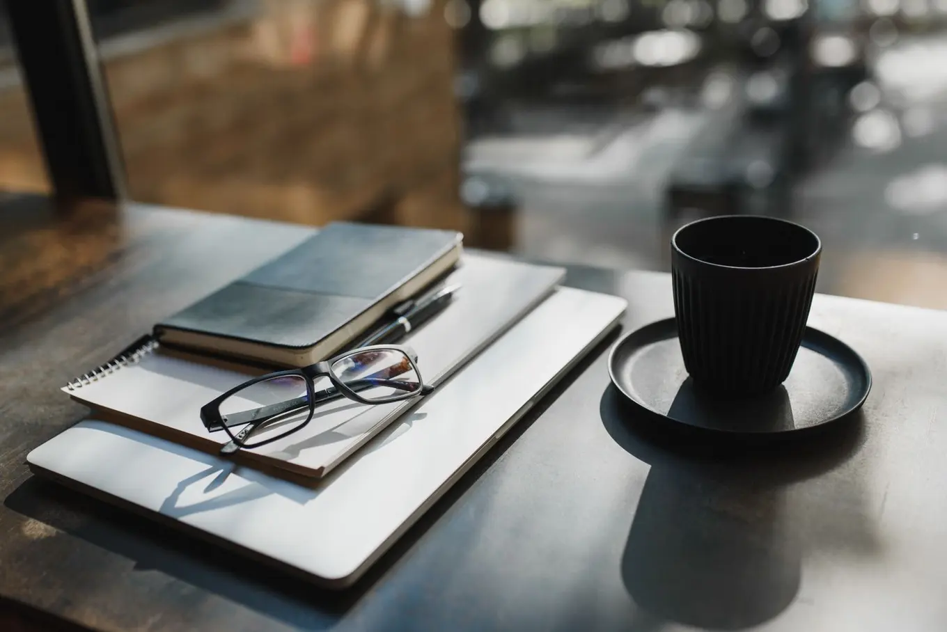 A notebook, a pair of glasses and a black coffee cup on a table by a window.