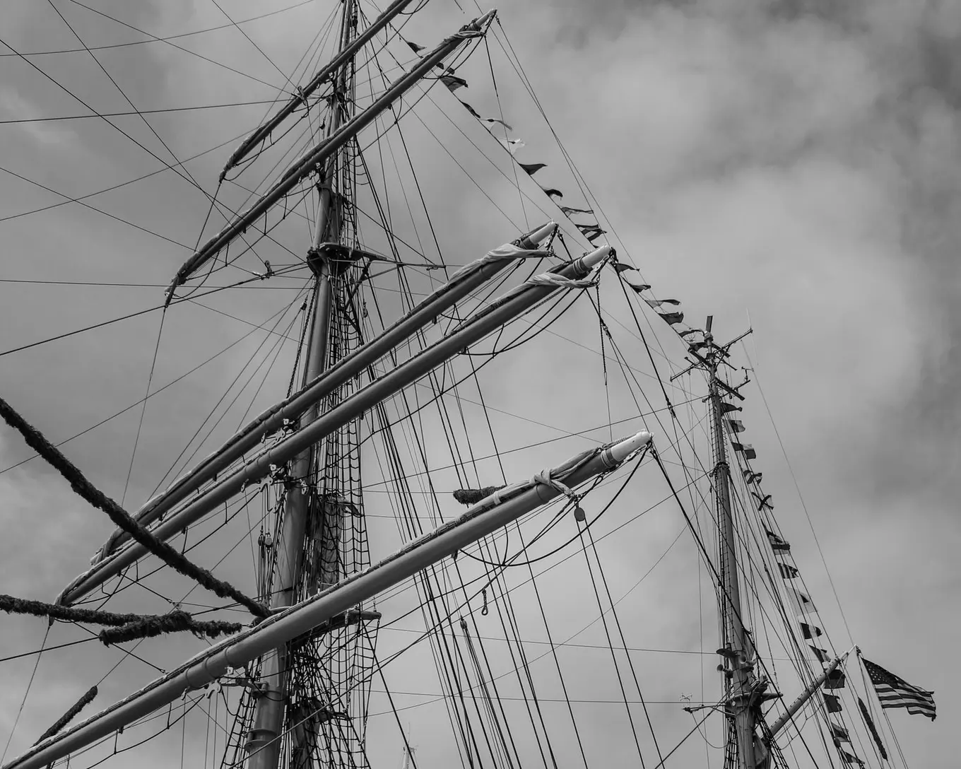 Black and white photo of a tall ship’s masts and rigging against a cloudy sky.