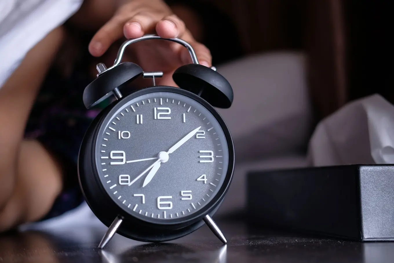 A hand reaching out from bed to switch off a black alarm clock.