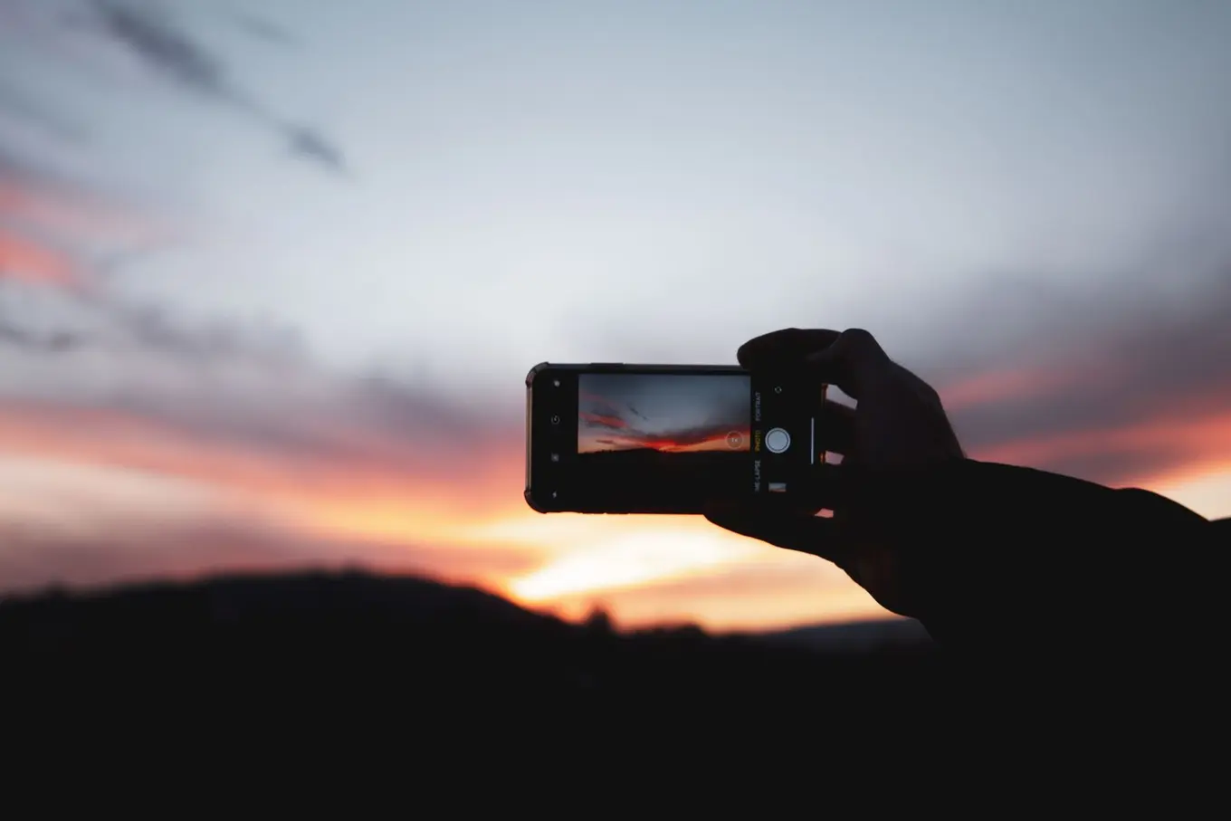 A hand holding up a phone to photograph a pink sky over a dark hill.