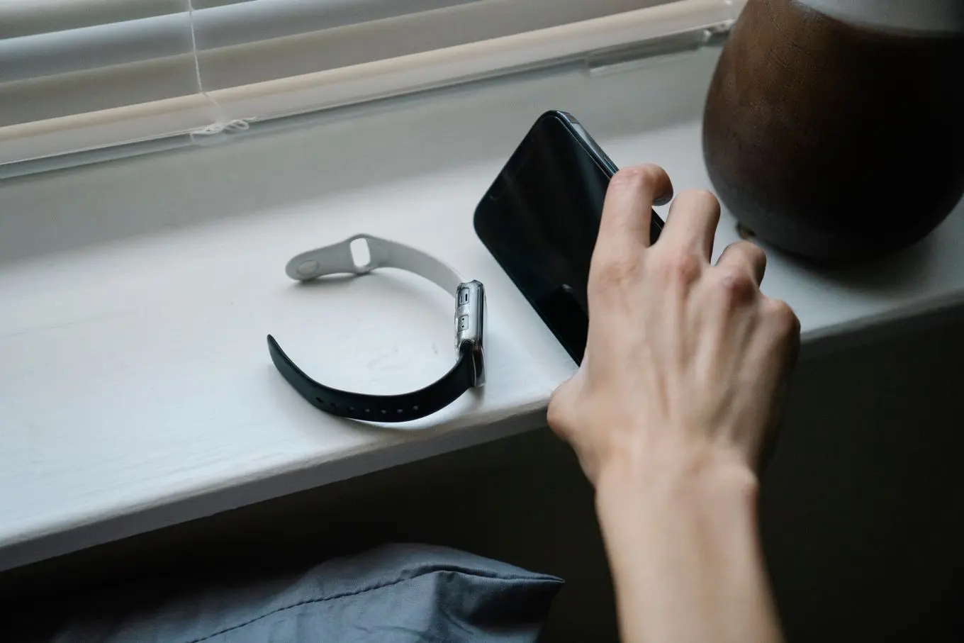 A hand reaching for a phone on a bedroom windowsill next to a smartwatch, a pillow just visible below