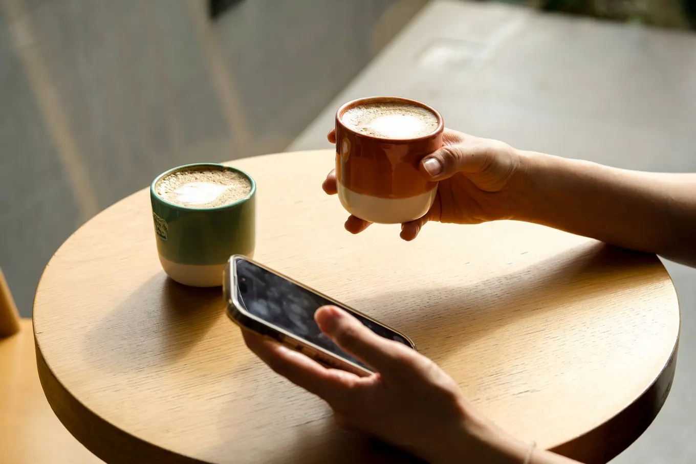 A hand holding a coffee cup while the other scrolls a phone on a round wooden table.
